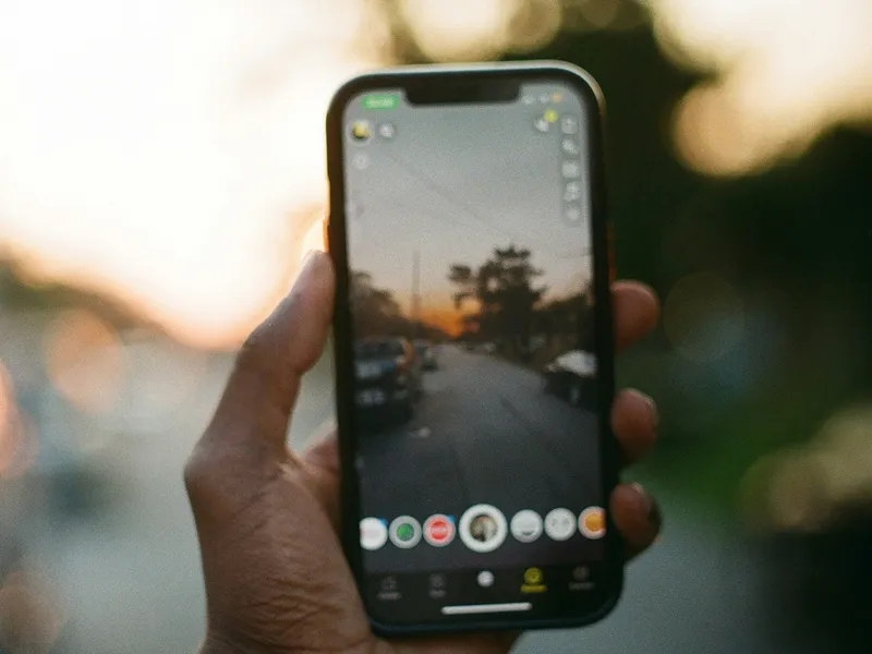 Person holding a smartphone vertically, capturing a photo of a sunset-lit street with trees and parked cars, as seen through the phone's camera interface.