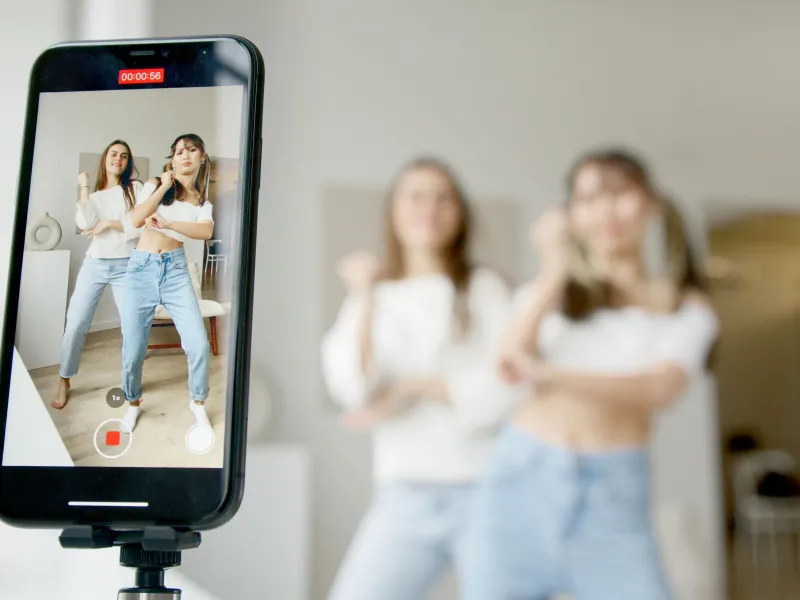 Two young women dance in front of a smartphone recording their choreography. Two other women watch in the background.