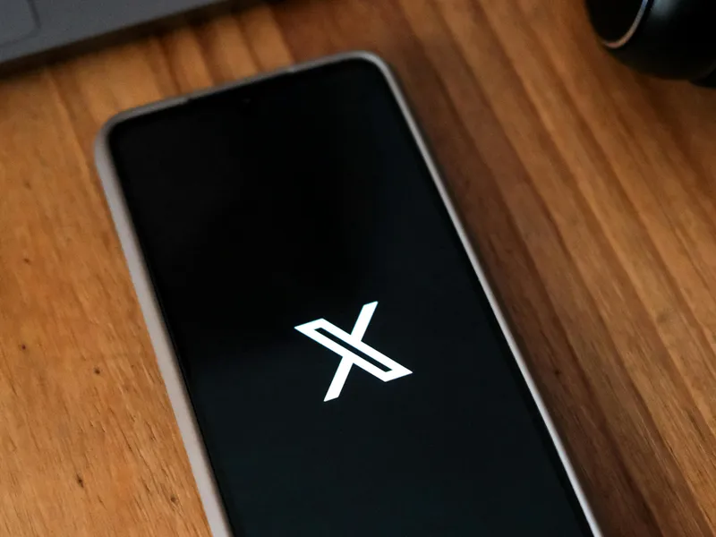 Smartphone displaying the X logo of the rebranded Twitter app on a wooden desk beside a laptop and headphones