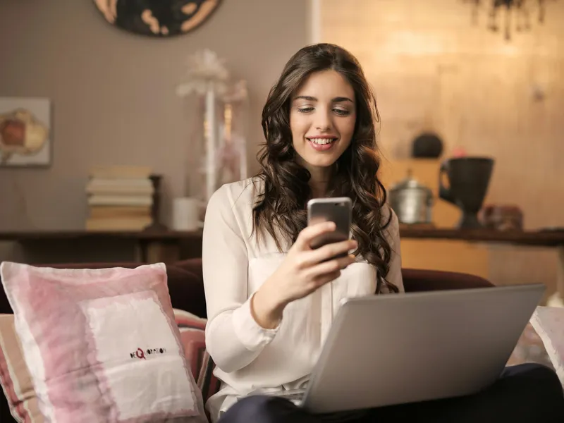 Smiling woman using smartphone while working on a laptop at home, representing social media engagement or remote digital communication
