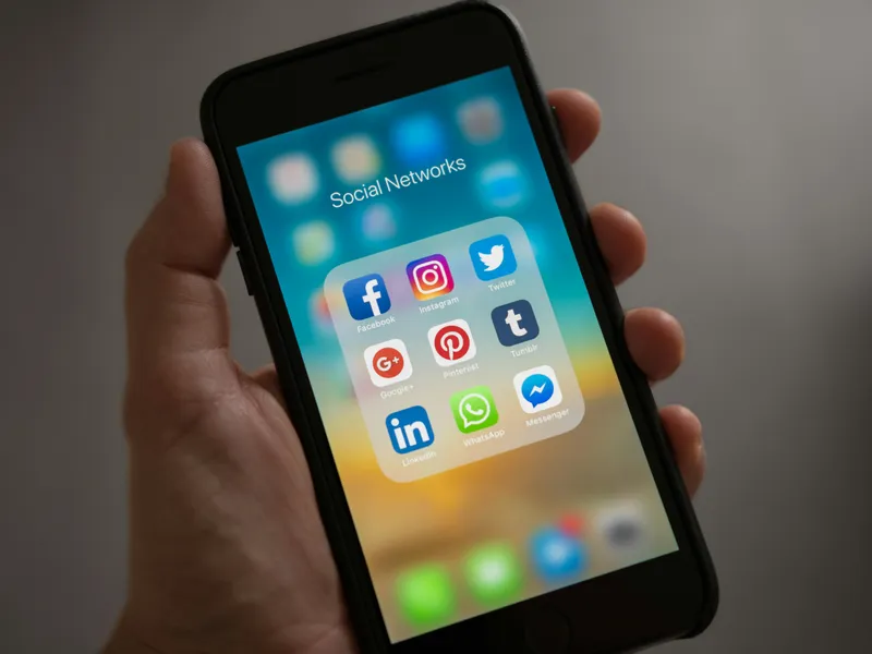 Person holding smartphone displaying social media folder with popular app icons including Facebook, Instagram, Twitter, LinkedIn, WhatsApp, and Messenger on the home screen
