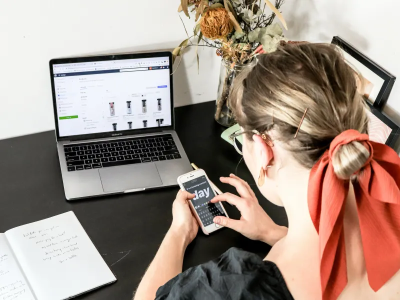 Person working at desk, using laptop and phone to manage online store.